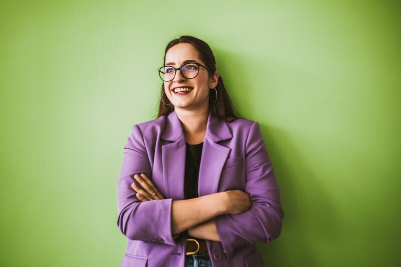 Eine Frau in einem lila Blazer posiert an einer grünen Wand und lächelt.